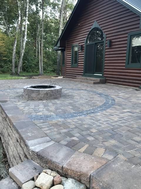 Brick patio with a fire pit, steps up to a log cabin with green trim.