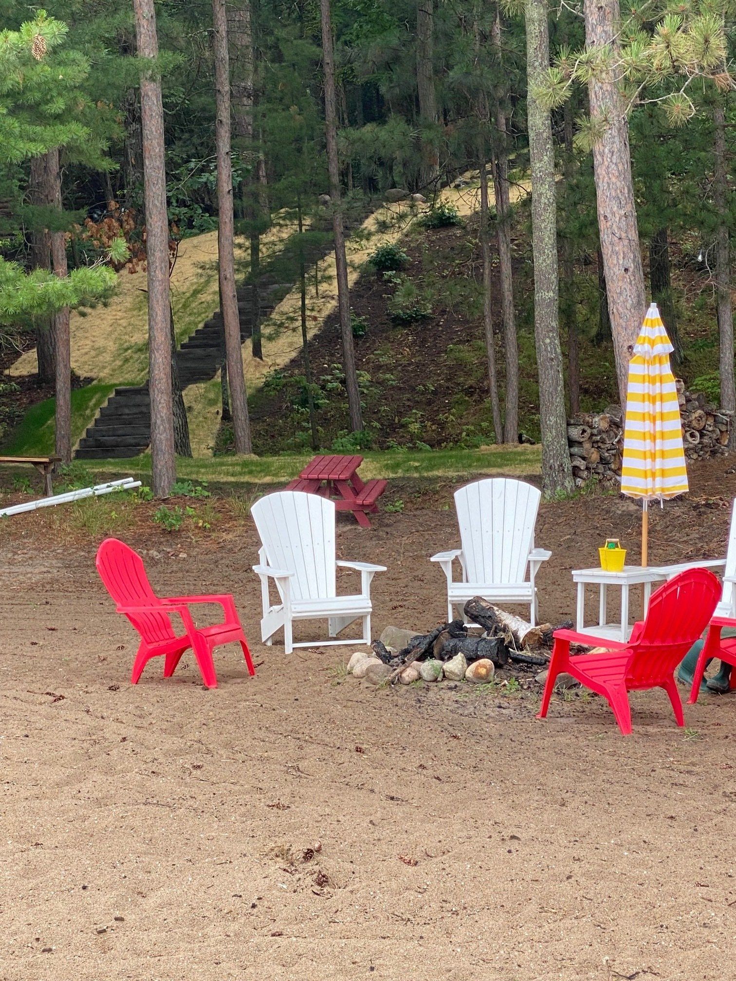 Beach scene: red and white Adirondack chairs around a fire pit, yellow umbrella, trees, stairs leading up a hill.