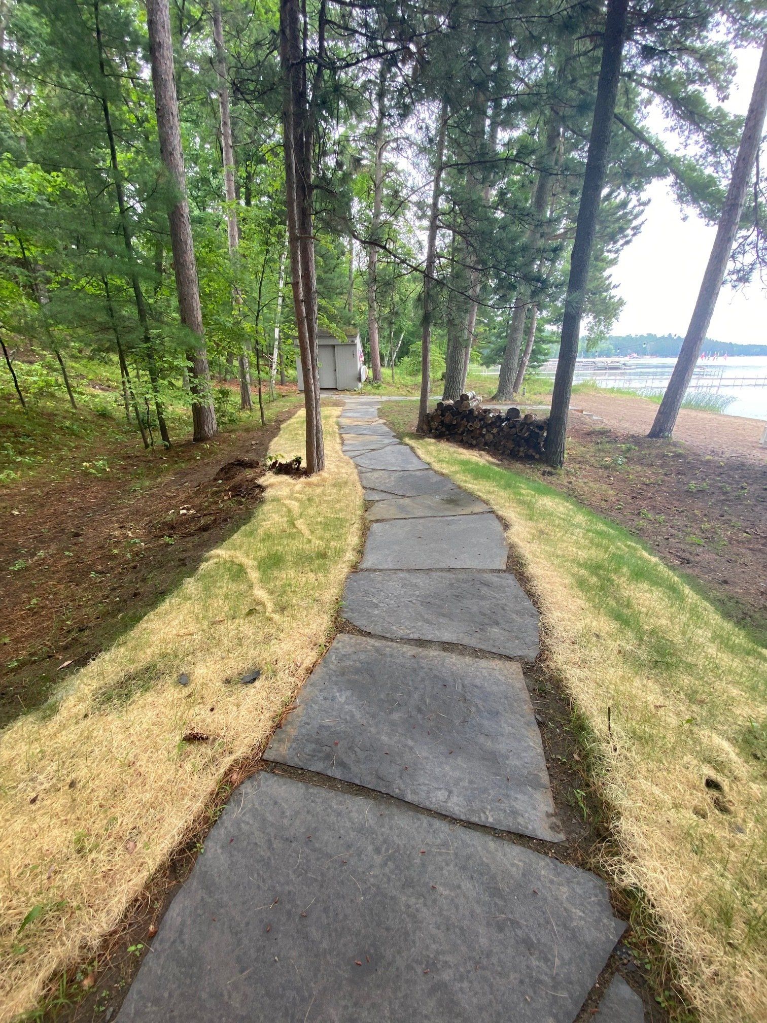 Stone path through a wooded area leading towards a lake; green trees surround path with some dry grass.