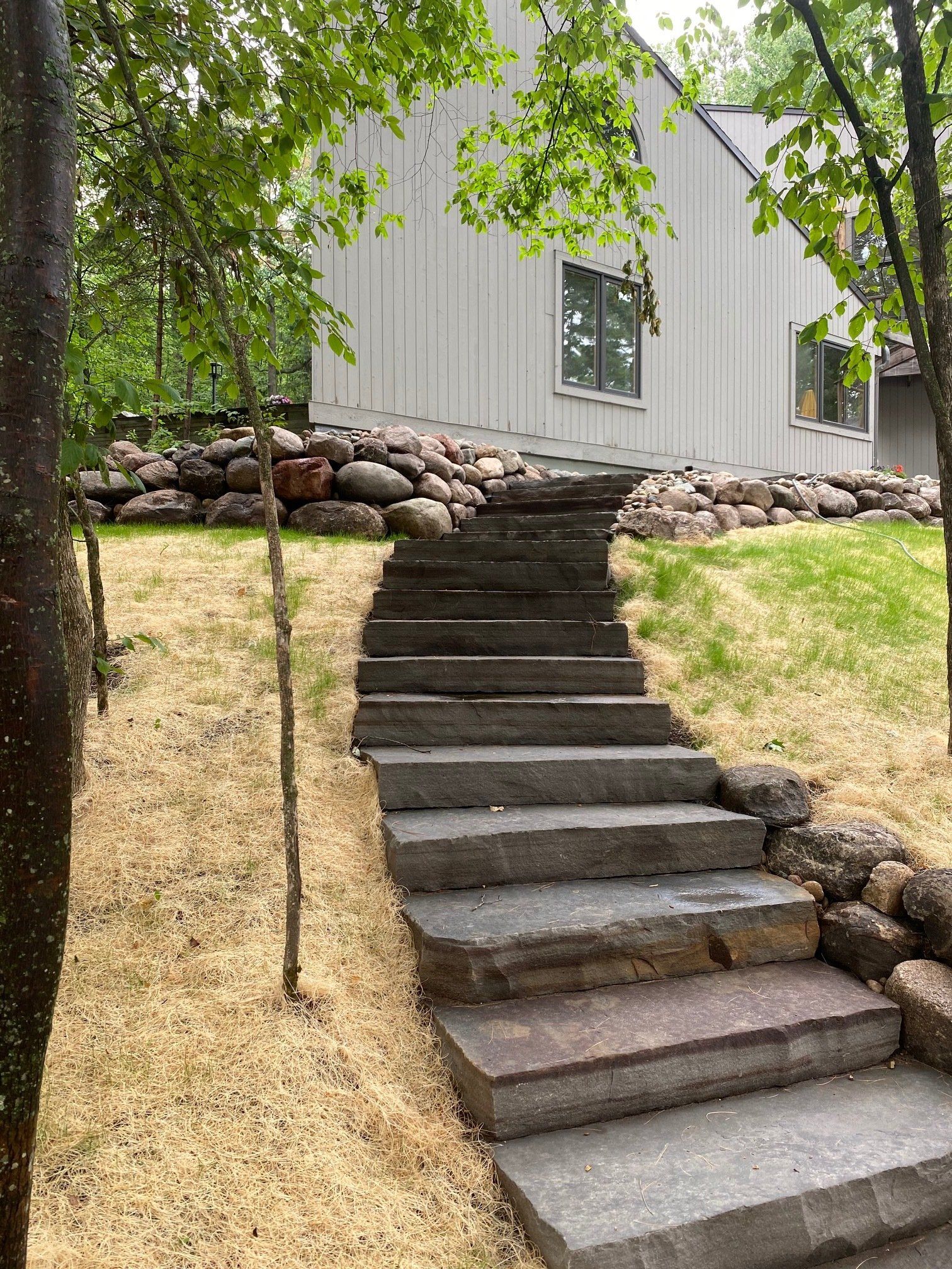 Stone steps leading up to a light grey house with a rock retaining wall and trees.