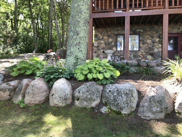 Large rocks form a border around a garden with hostas near a stone house with a wooden balcony.
