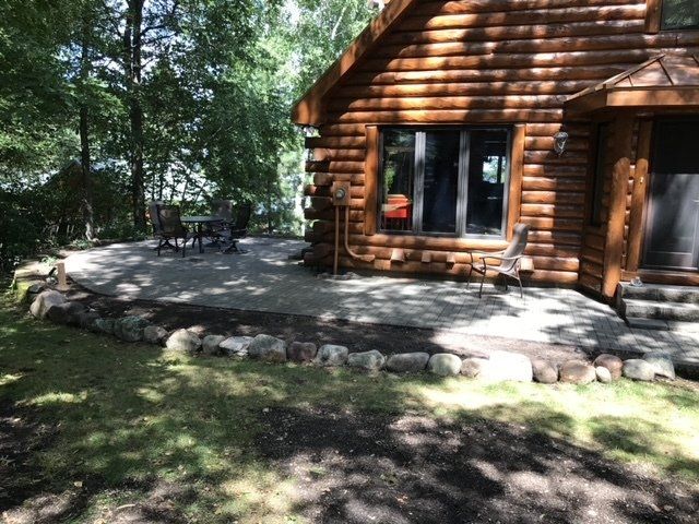 A log cabin with a stone patio, trees in the background, and a table and chairs.
