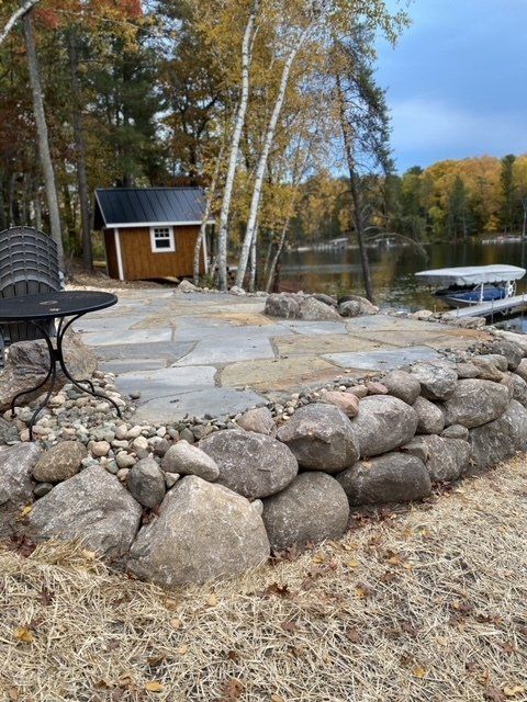 Stone patio with rock wall by lake; small brown shed in the background.