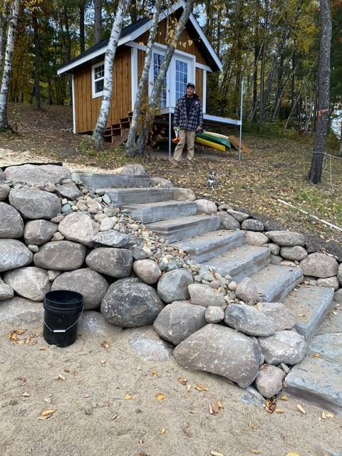 Man stands by a cabin with stone steps leading down to a sandy area, with canoes and firewood nearby.