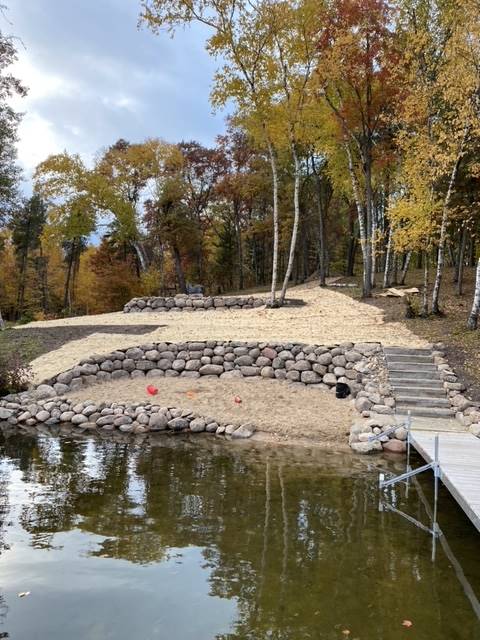Lakefront with sandy beach area, stone retaining walls, steps, and dock, fall foliage in the background.