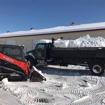Snow removal with a Kubota skid steer loading a truck with snow in front of a building on a sunny day.