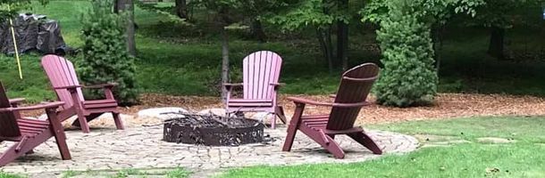 A fire pit surrounded by Adirondack chairs in a grassy backyard. Green trees in the background.
