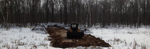 A bulldozer digging a ditch in a snowy field in front of a forest of leafless trees.