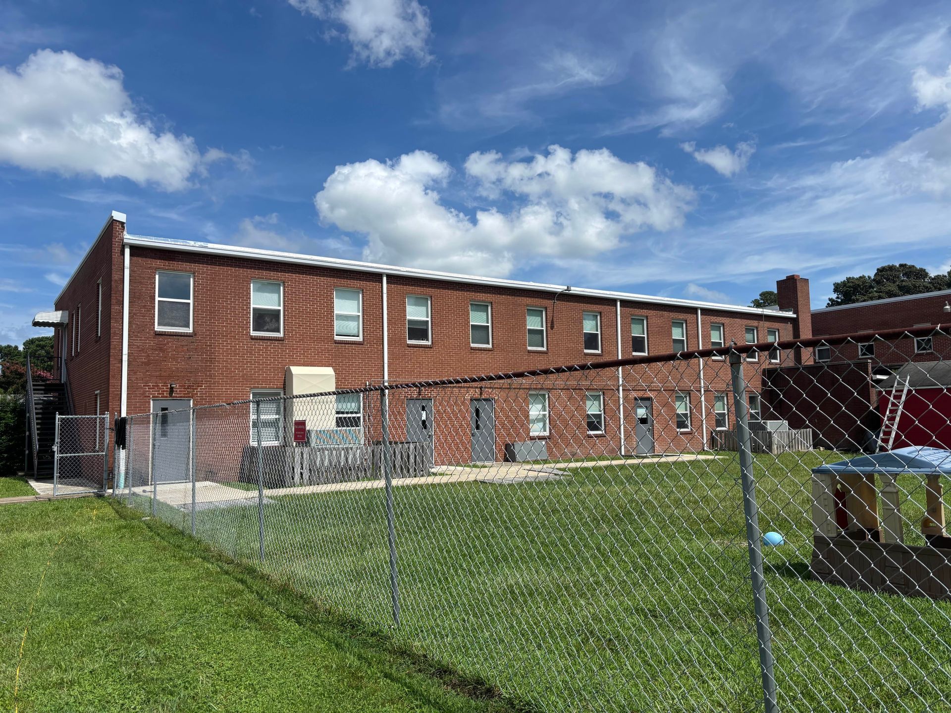 Brick building behind a chain-link fence on a grassy lot under a blue, cloudy sky