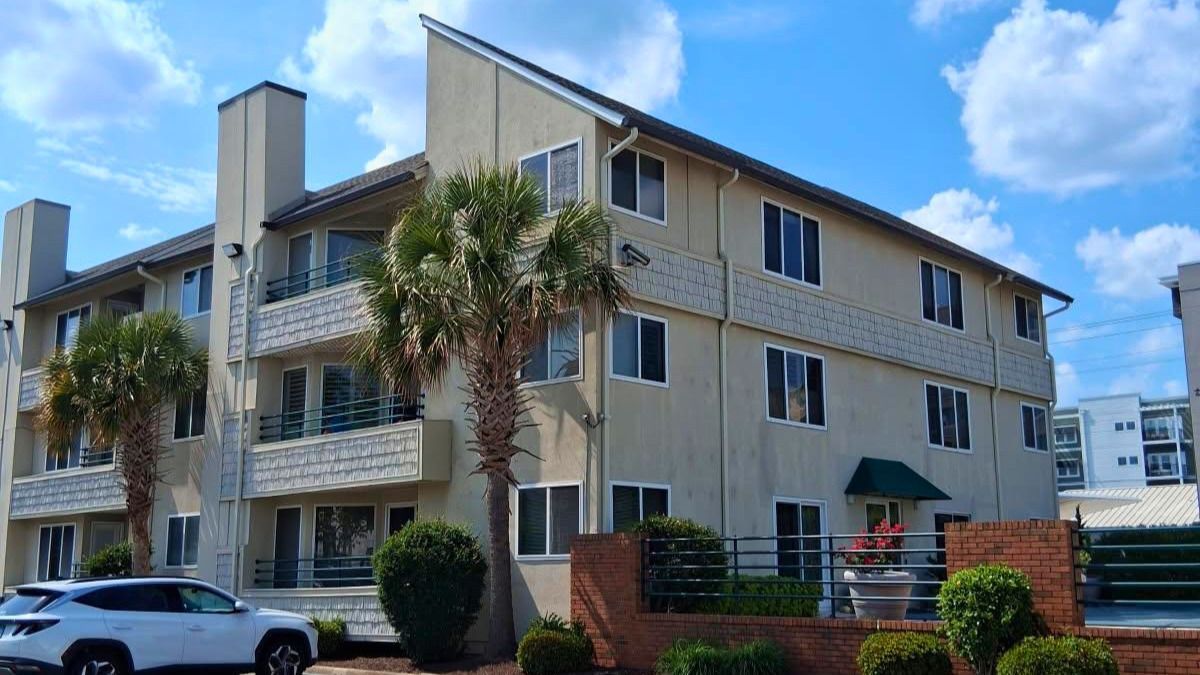 Three-story beige apartment building with balconies, palm trees, and parked cars under a blue sky