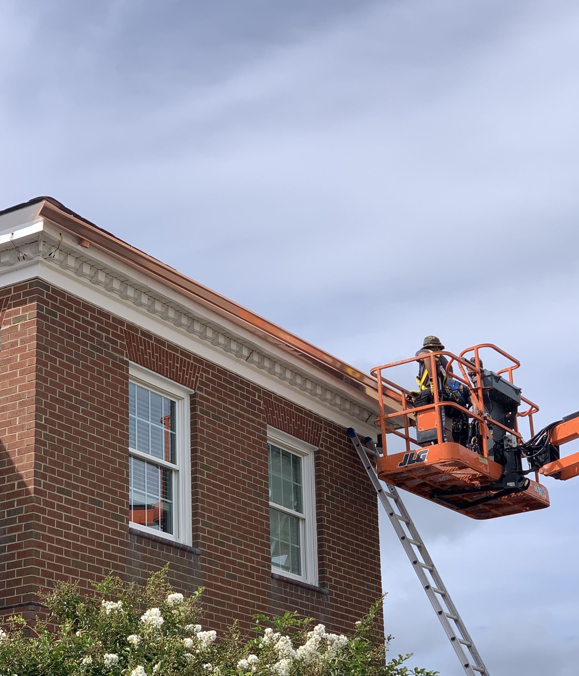 Worker in an orange lift inspecting a brick building roof near a ladder under a cloudy sky