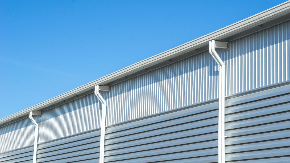 Metal warehouse siding with white gutters under a blue sky.