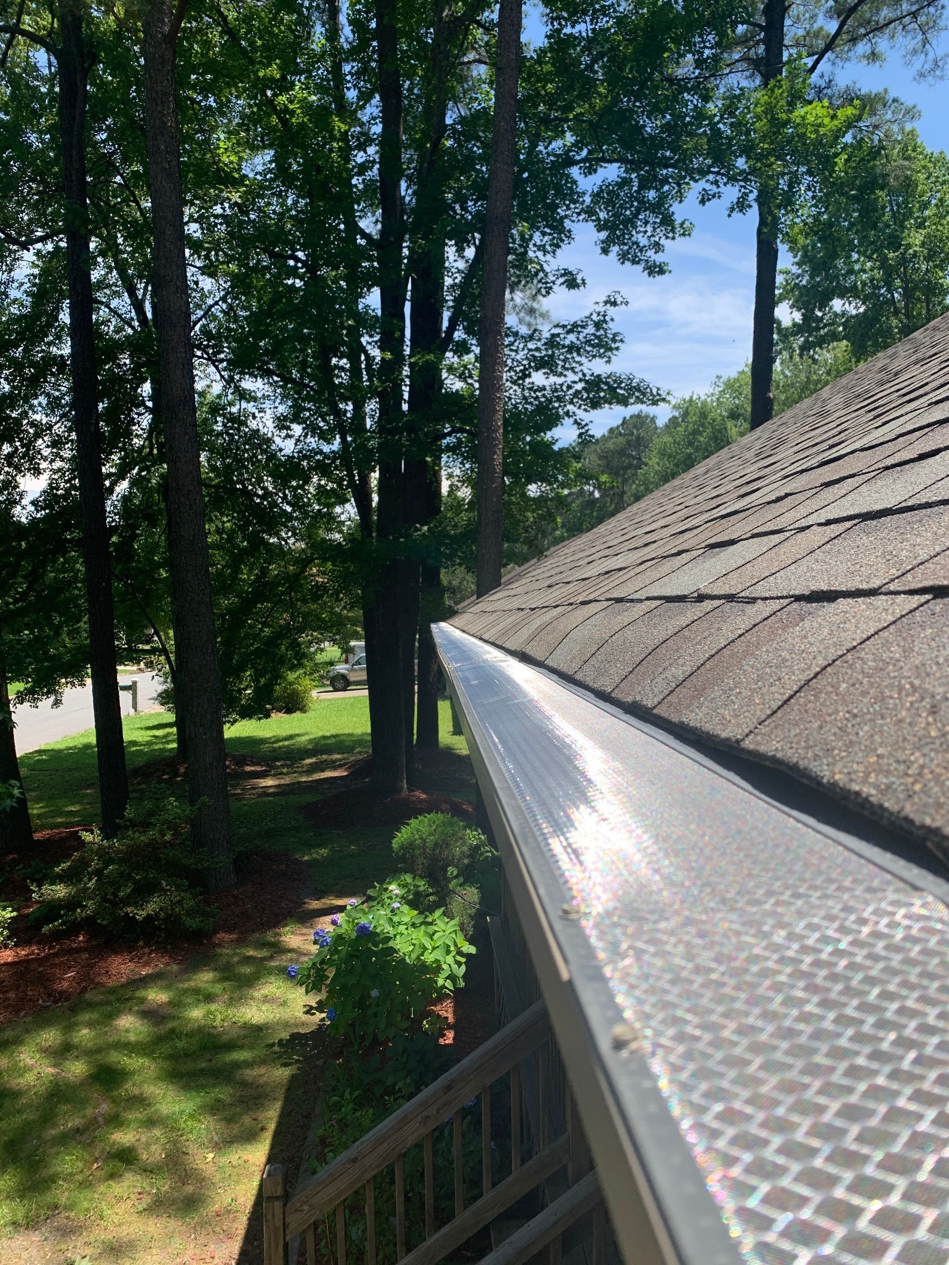 Gutter covered with a silver mesh sits against a gray shingled roof, surrounded by green trees and grass.