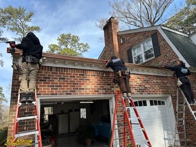 Three workers installing gutters on a brick house, using ladders and power tools. Sunny day.