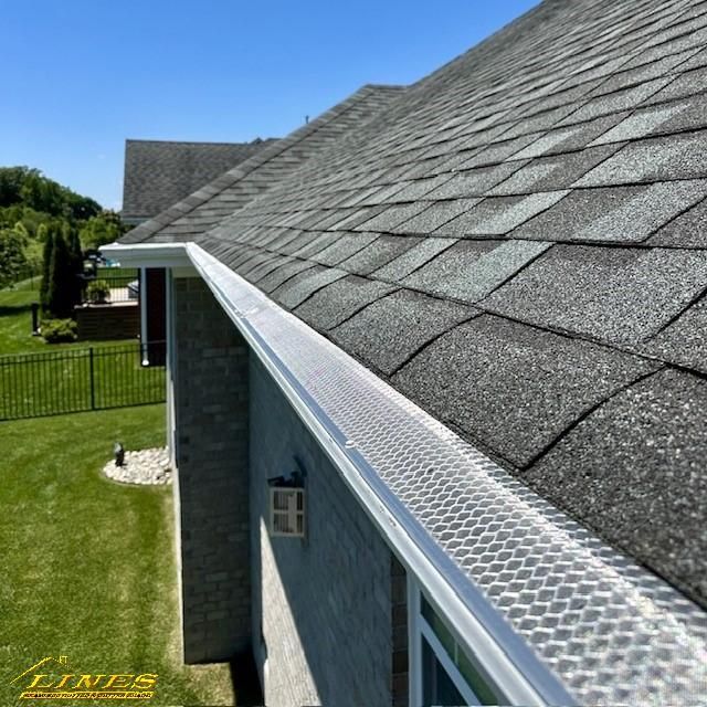 Gutter with mesh covering installed along a gray shingled roof on a sunny day.