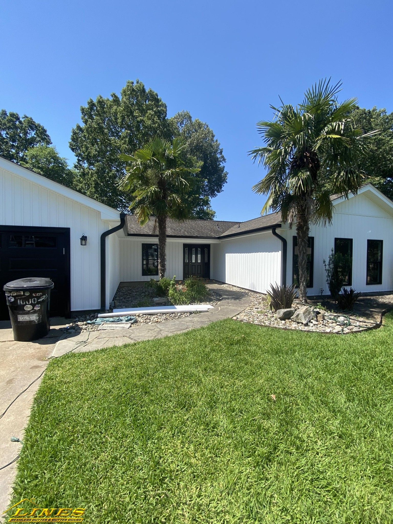 White house with black trim and garage; green lawn, trees, and clear blue sky.