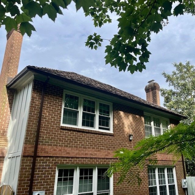 Brick house with white-trimmed windows, a chimney, and a dark roof, under a cloudy sky, viewed through leaves.