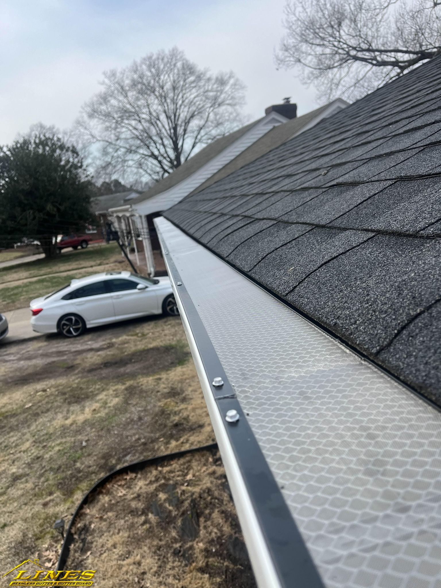Gutter with mesh guard along a dark shingle roof. A white car is parked in the background.