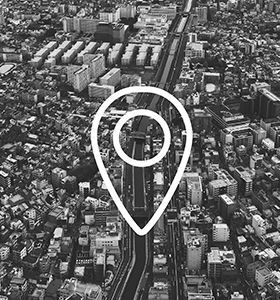 Black-and-white aerial city view with a large map pin icon centered over a road or rail corridor.