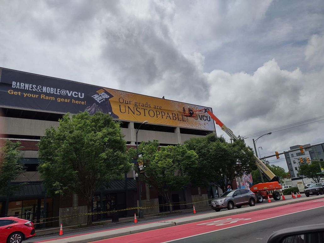 Large building with a banner, trees in front, and cars along a street under a cloudy sky.