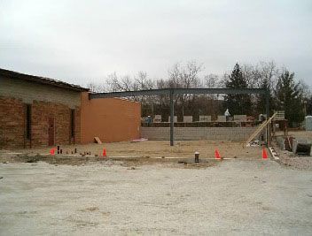 Construction site with a partially built steel frame structure attached to a brick wall under a cloudy sky.
