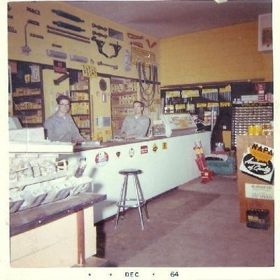 Two store employees stand behind a service counter at a NAPA Auto Parts store, decorated with wall-mounted auto tools, 1964.