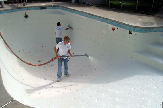 Two people cleaning an empty swimming pool with white walls, one spraying with a hose, the other scrubbing.