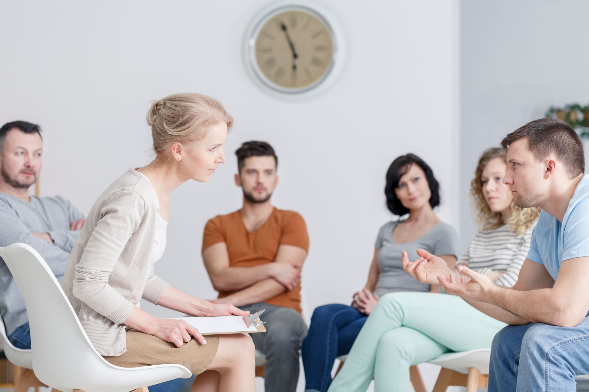 Small group in a therapy circle, seated in a bright room with a wall clock, one woman speaking to others.