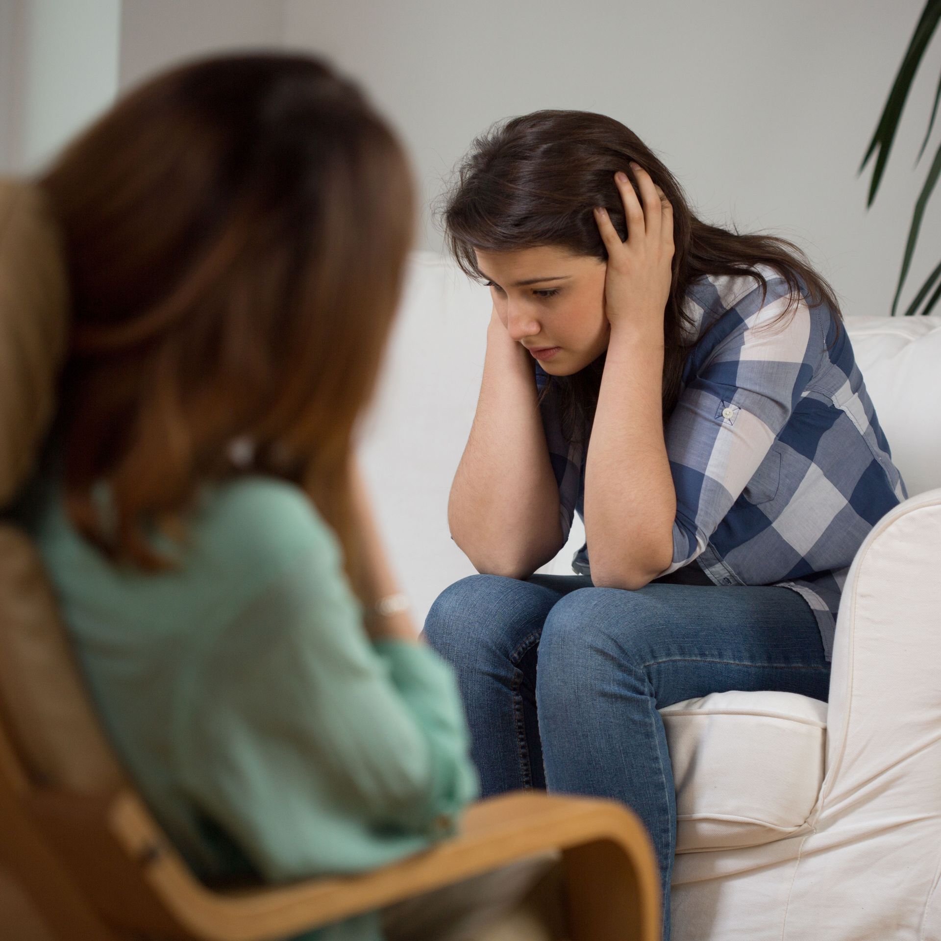 Woman sits hunched on a couch, holding her head while a blurred listener faces her.