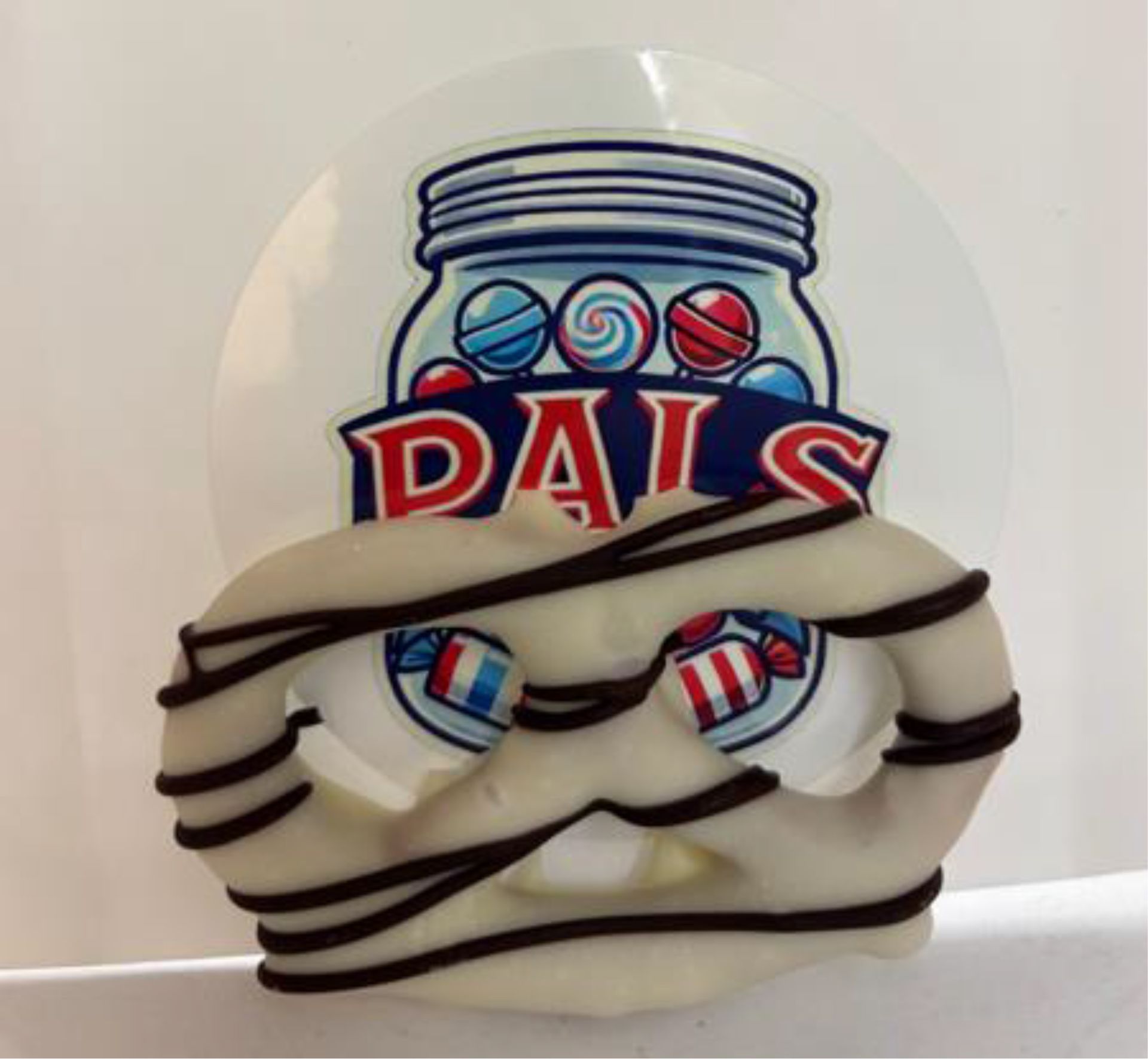 White chocolate-covered pretzel with chocolate drizzle, set in front of a Pals candy jar logo.