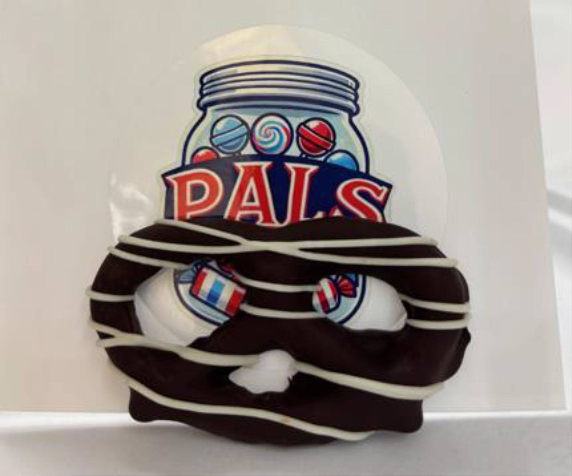 Chocolate-covered pretzel with candy eyeballs, white drizzle, and PALS label featuring a jar of candies.