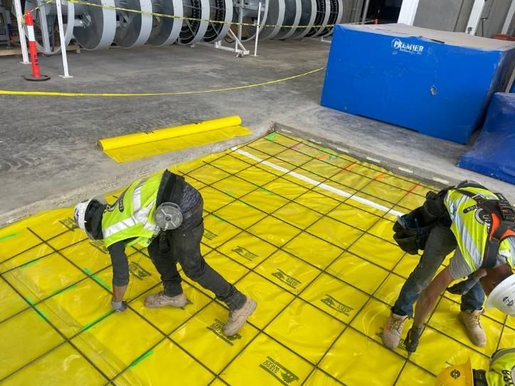 Two construction workers are working on a yellow tarp on the ground.