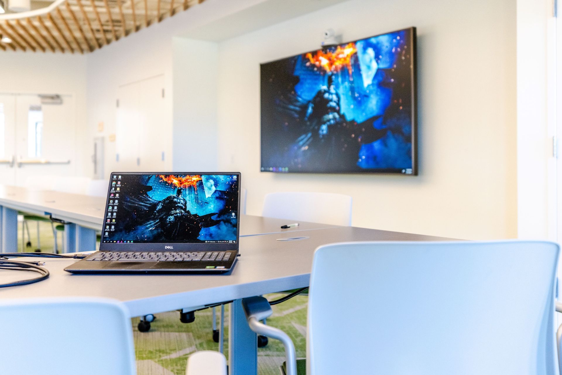 Laptop on a table in a bright office lounge with a wall-mounted display showing the same screen