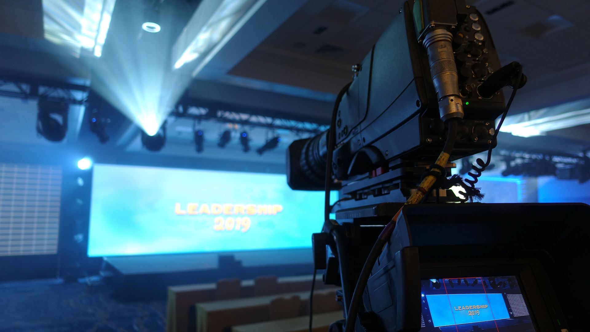 Camera in a blue-lit TV studio aimed at a large glowing screen and stage lights.