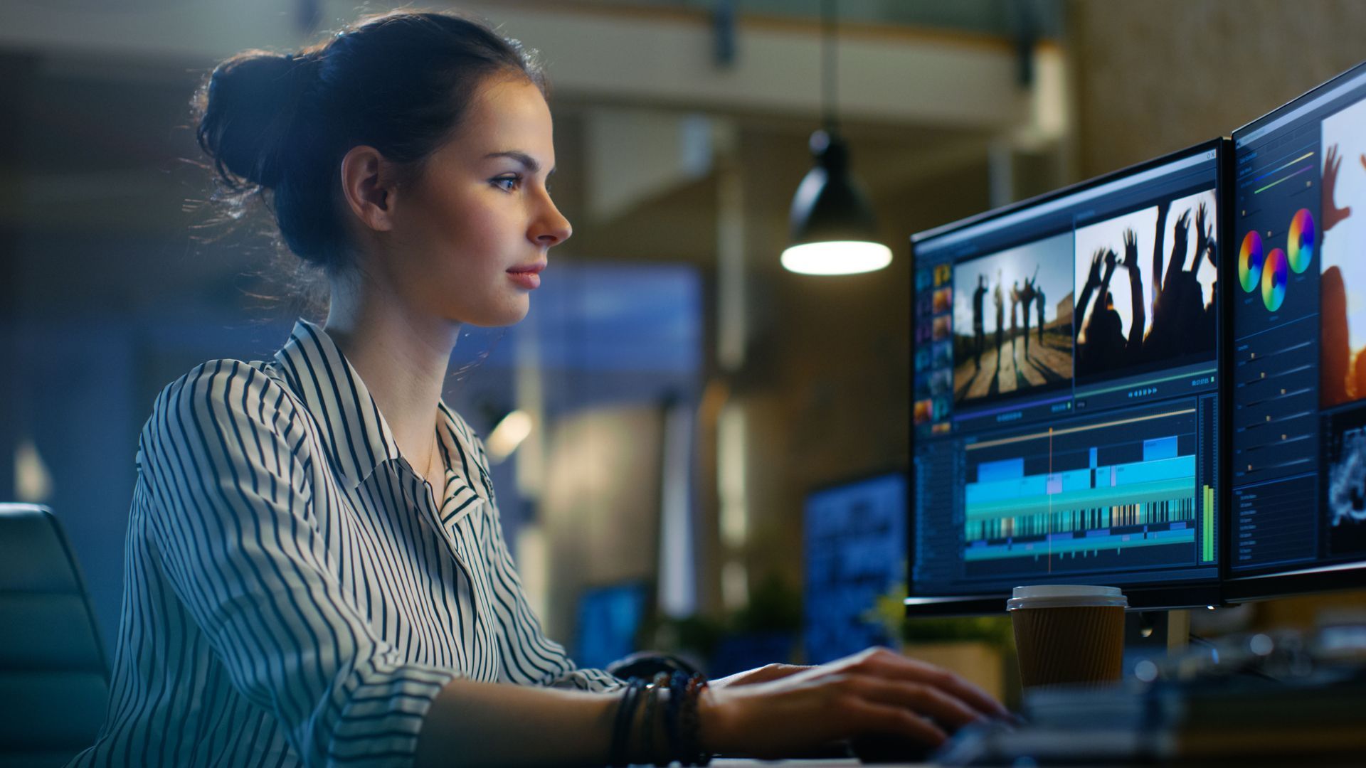 Person working at a desk with dual monitors in a dimly lit office, focused on video editing.