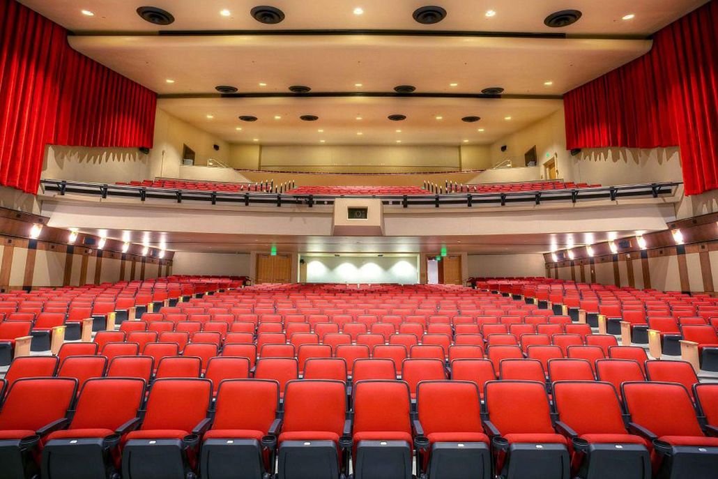 Empty auditorium with red seats, stage, and balcony under warm ceiling lights