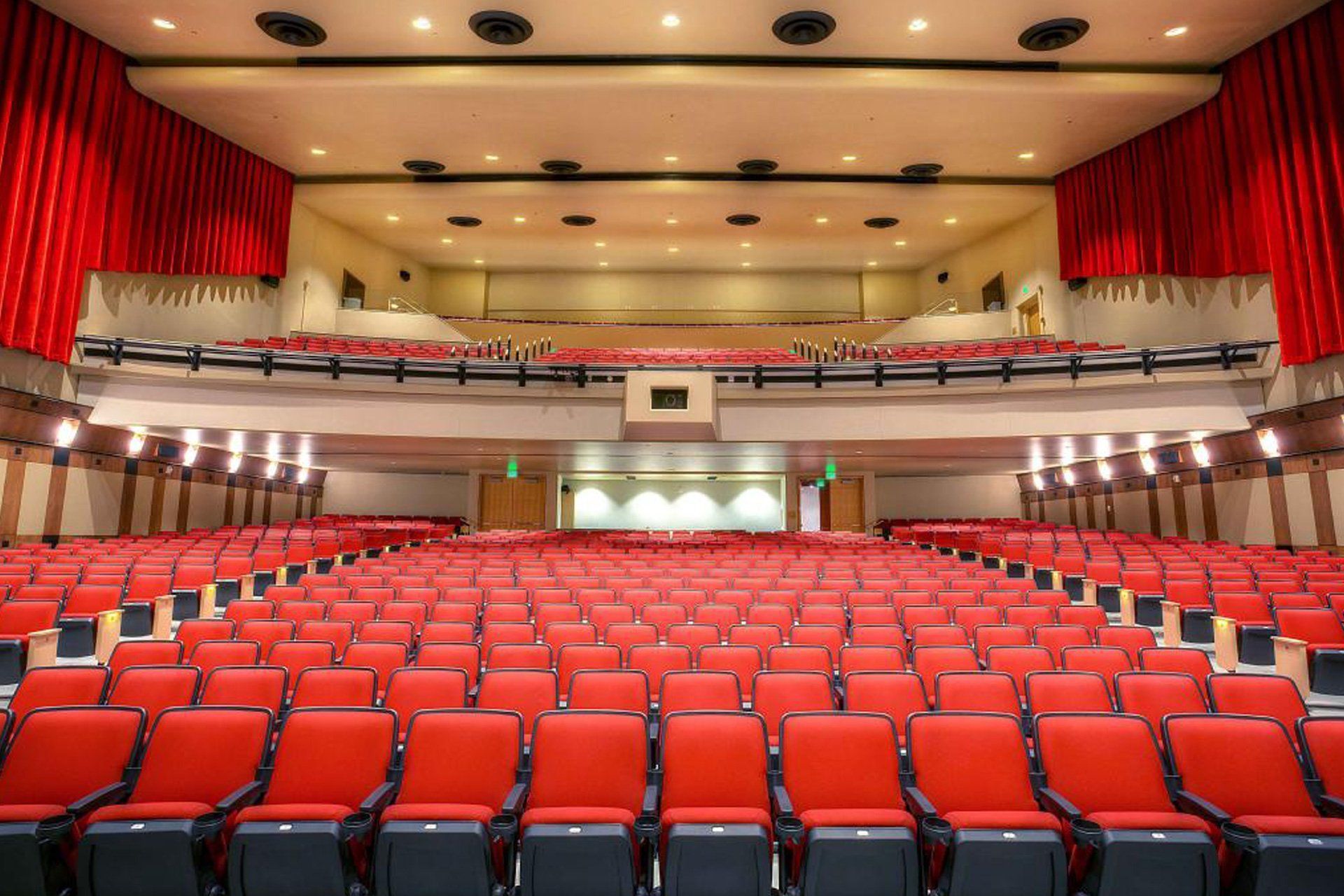 Empty auditorium with red seats, stage, and balcony under warm ceiling lights