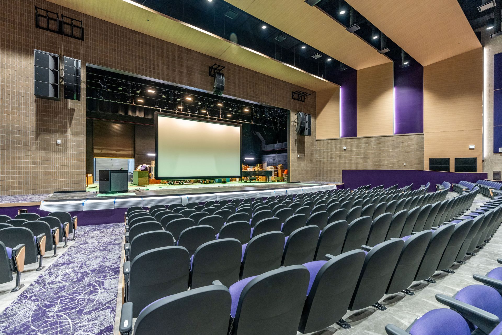 Empty conference hall with rows of black chairs facing a large stage and screen in a modern auditorium