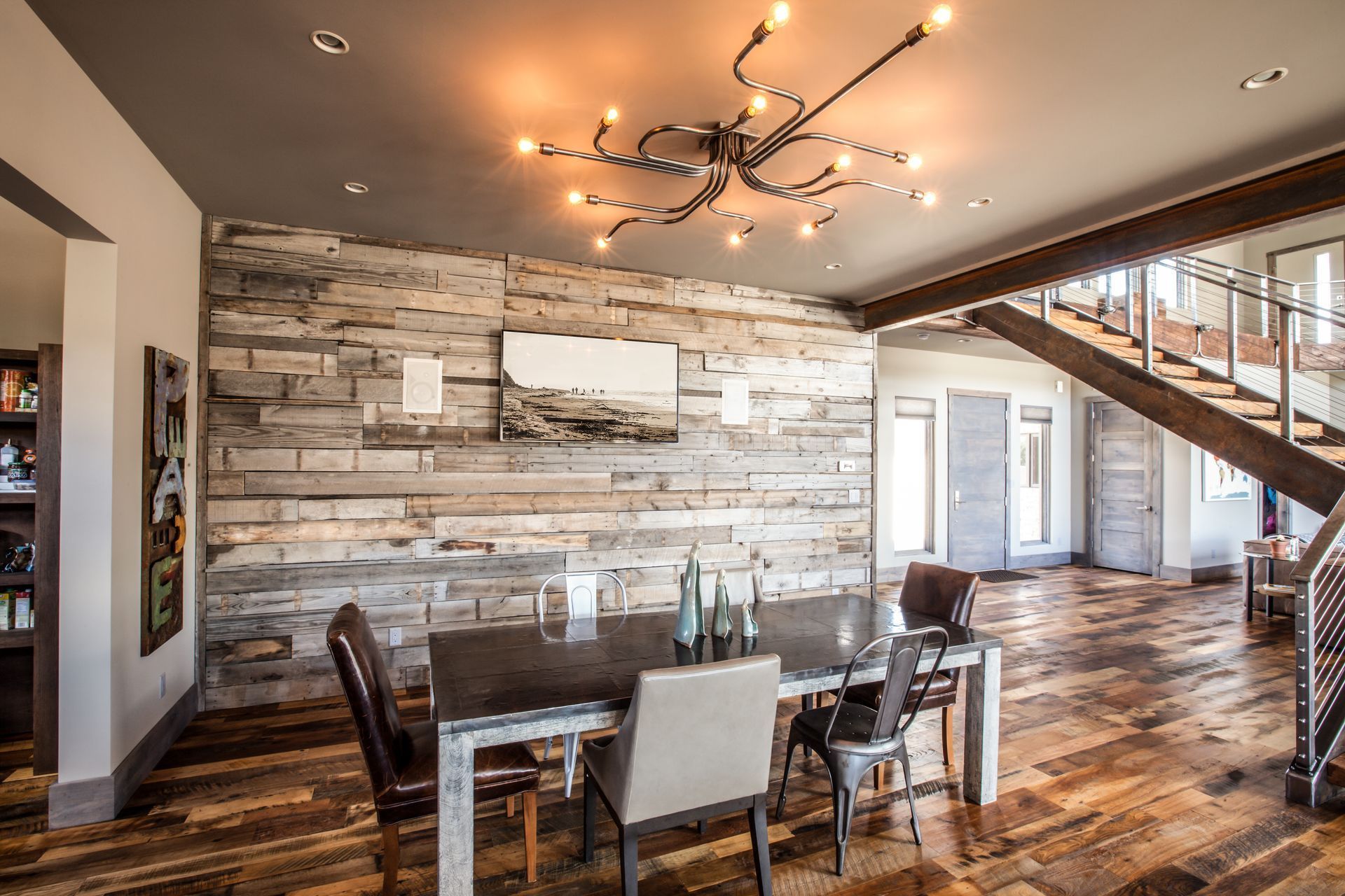 Modern dining room with rustic wood wall, glass table, leather chairs, and staircase by large windows
