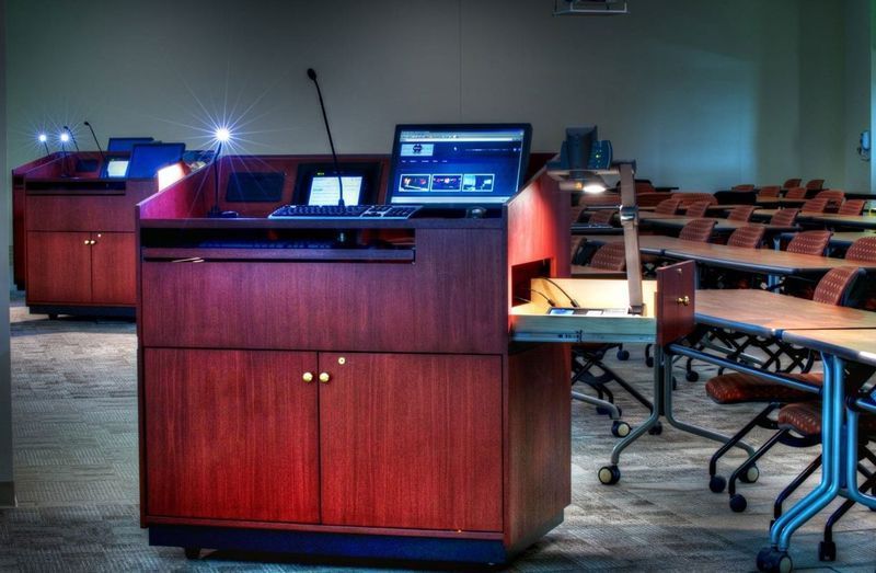 Wooden lecterns with monitors in a dim lecture hall with rows of chairs and tables