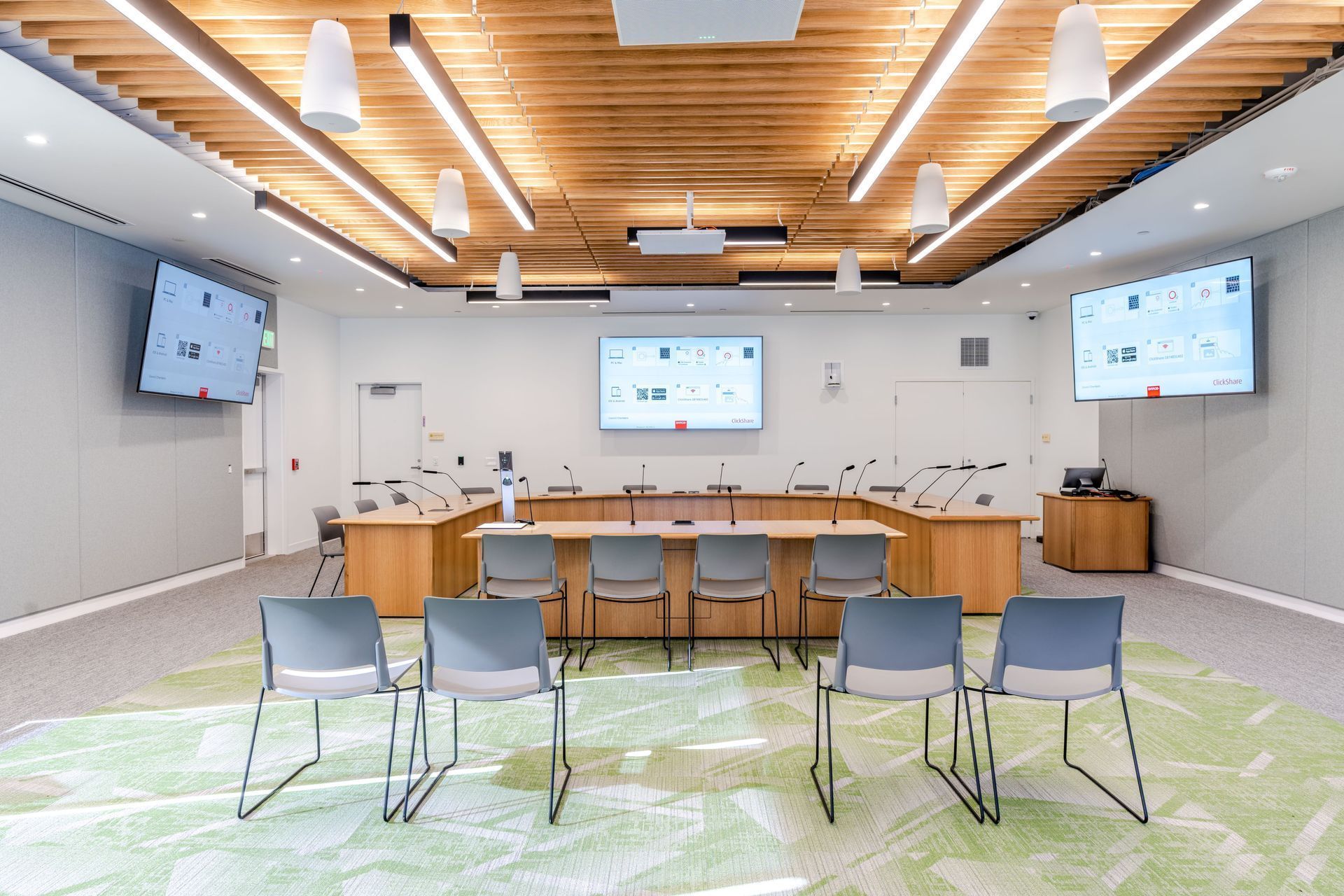 Modern conference room with rows of chairs, a long table, wall screens, and a wooden ceiling