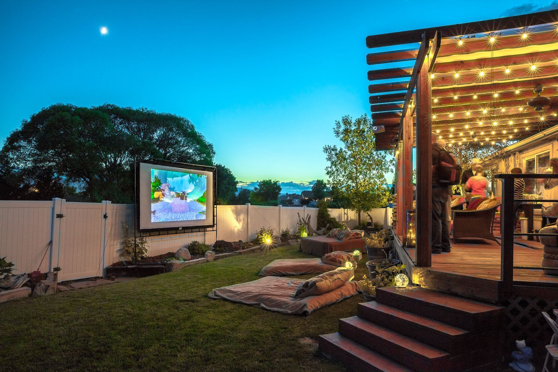 Backyard patio with string lights and outdoor screen beside a lawn at dusk