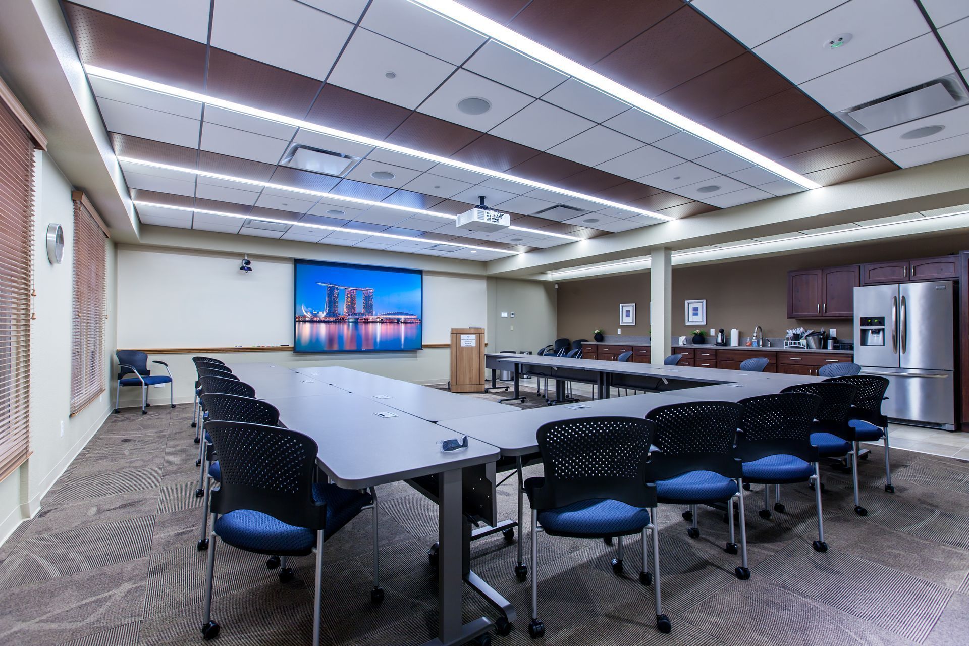 Modern conference room with U-shaped tables, black chairs, and a wall-mounted presentation screen