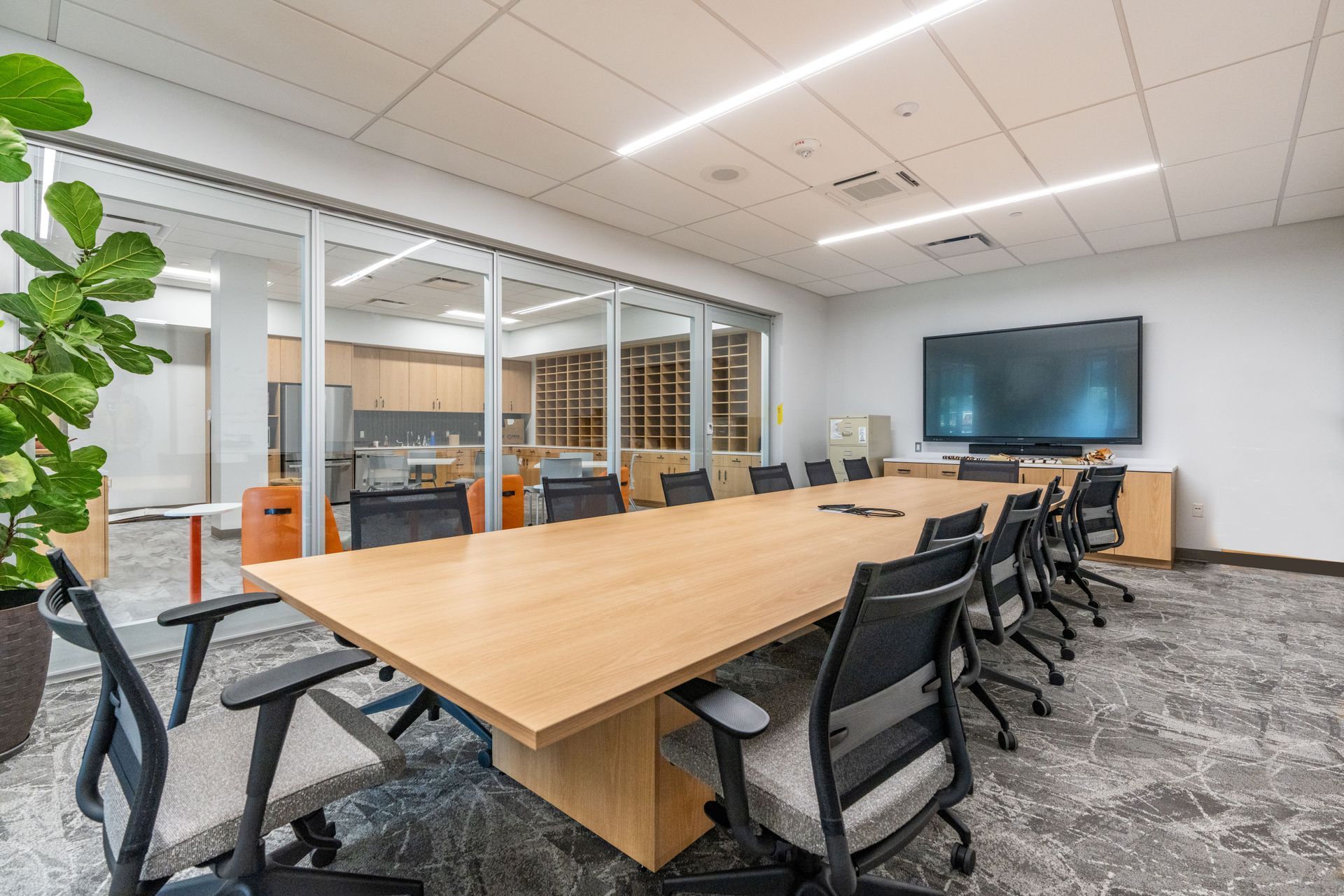 Modern conference room with a long wooden table, black chairs, glass walls, and a large wall-mounted screen