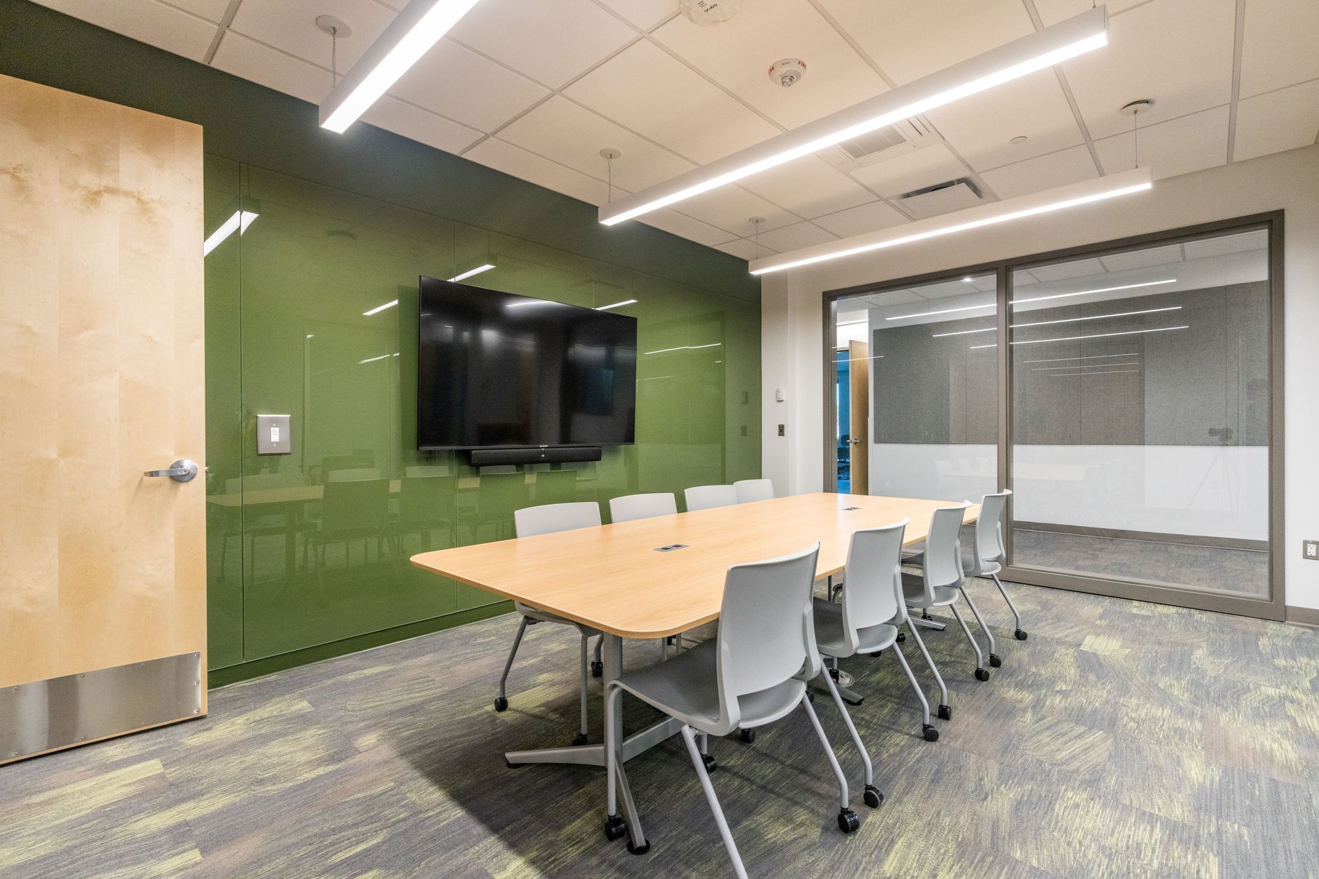 Modern conference room with long table, gray chairs, wall-mounted TV, and glass sliding doors.