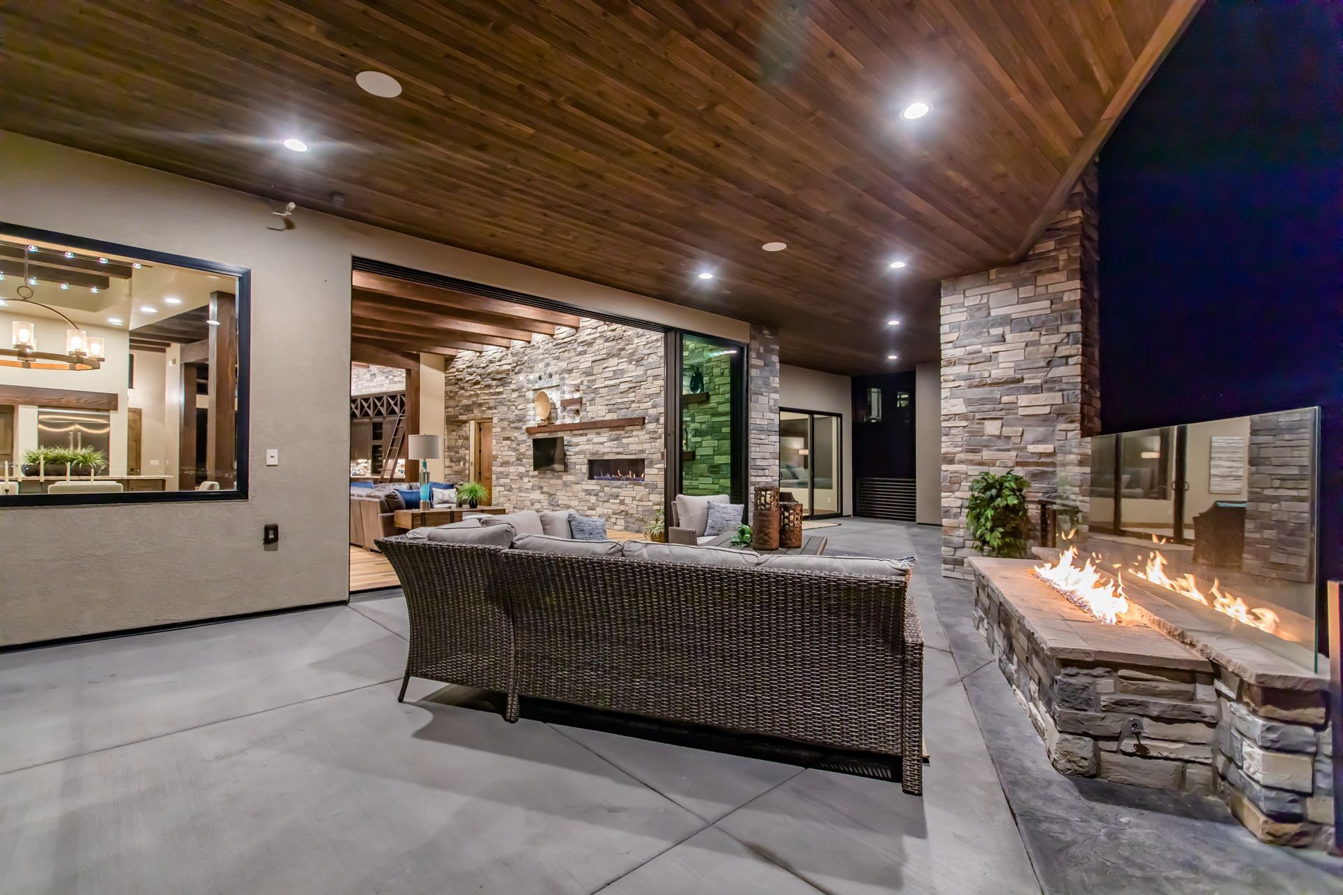 Covered patio with wicker seating, stone fireplace, and warm recessed lighting at night
