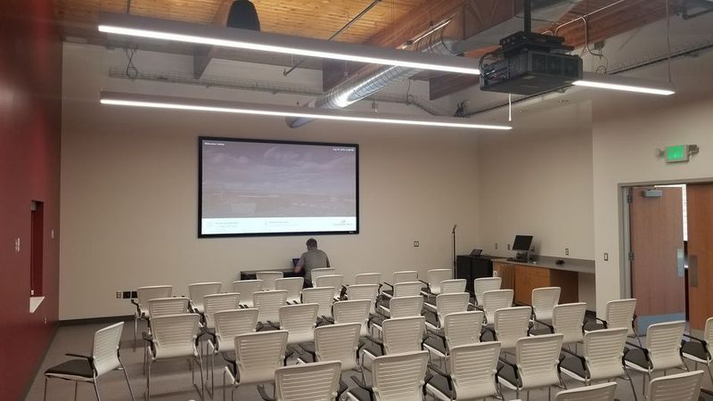 Empty classroom with rows of chairs, a projection screen, and a lectern near the front.