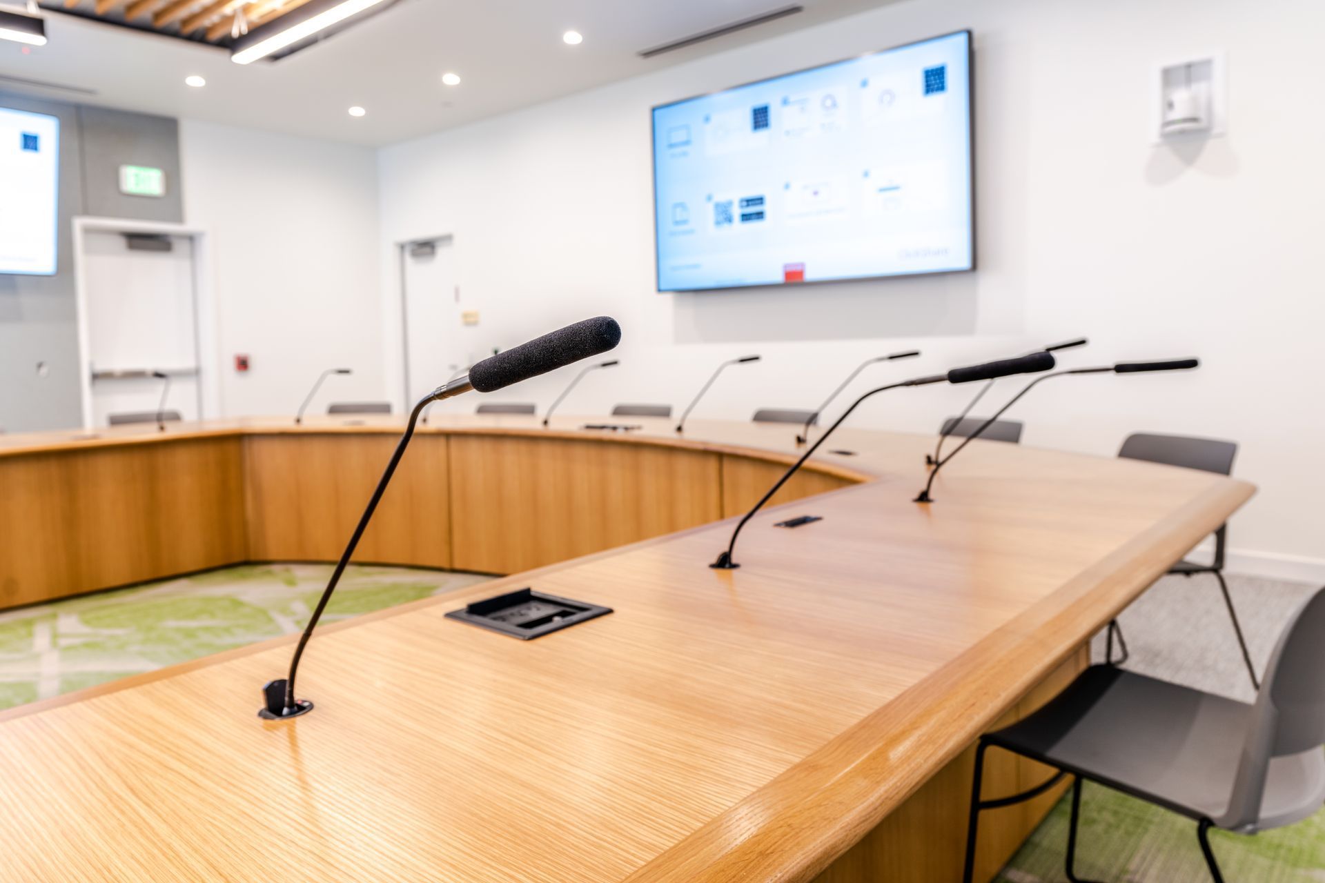 Modern conference room with a curved wooden table, gooseneck microphones, and a wall-mounted screen.