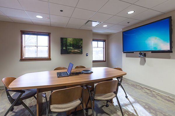 Conference room with wooden table, chairs, laptop, wall-mounted screen, and two windows