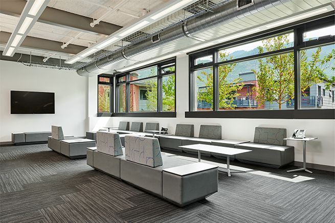 Modern office lounge with gray sofas, white tables, wall-mounted TV, and large windows.